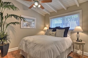 A bedroom with a bed, two bedside tables with lamps, a ceiling fan, a window with blinds, and a framed picture on the wall.