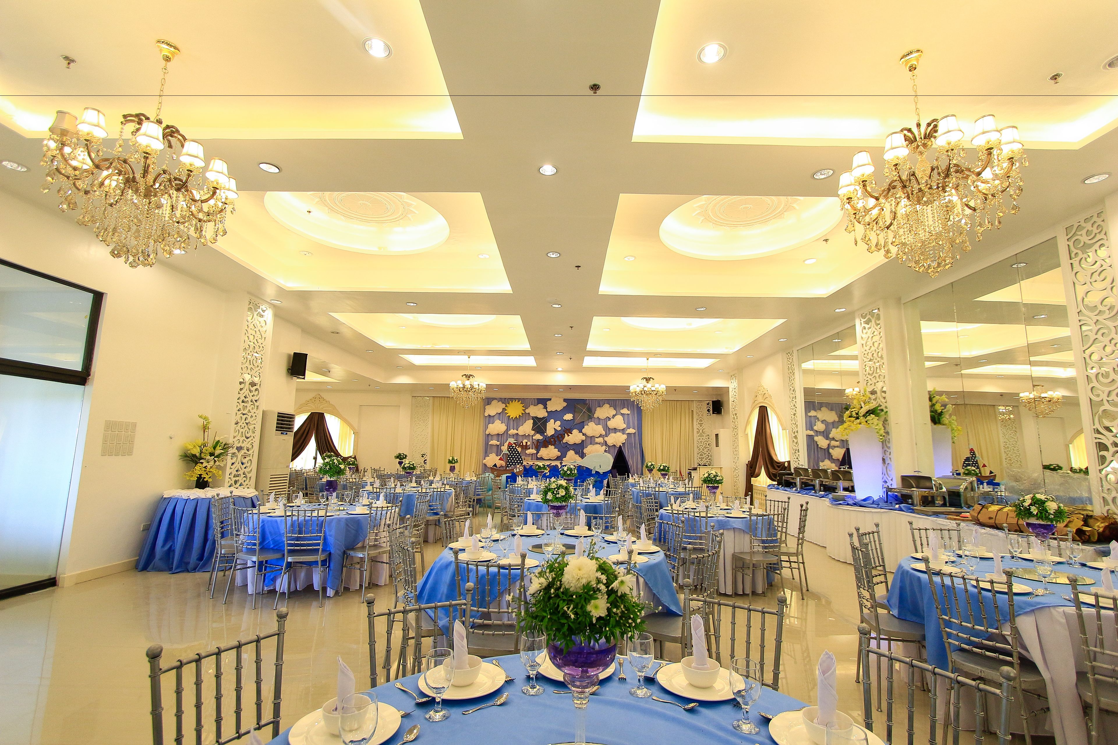 A banquet hall with tables set for a formal event, featuring chandeliers and a stage with floral decorations.