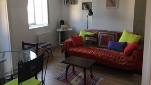 En stue med et glasbord, en sofa med farverige puder, en radiator, et ur og en reol.