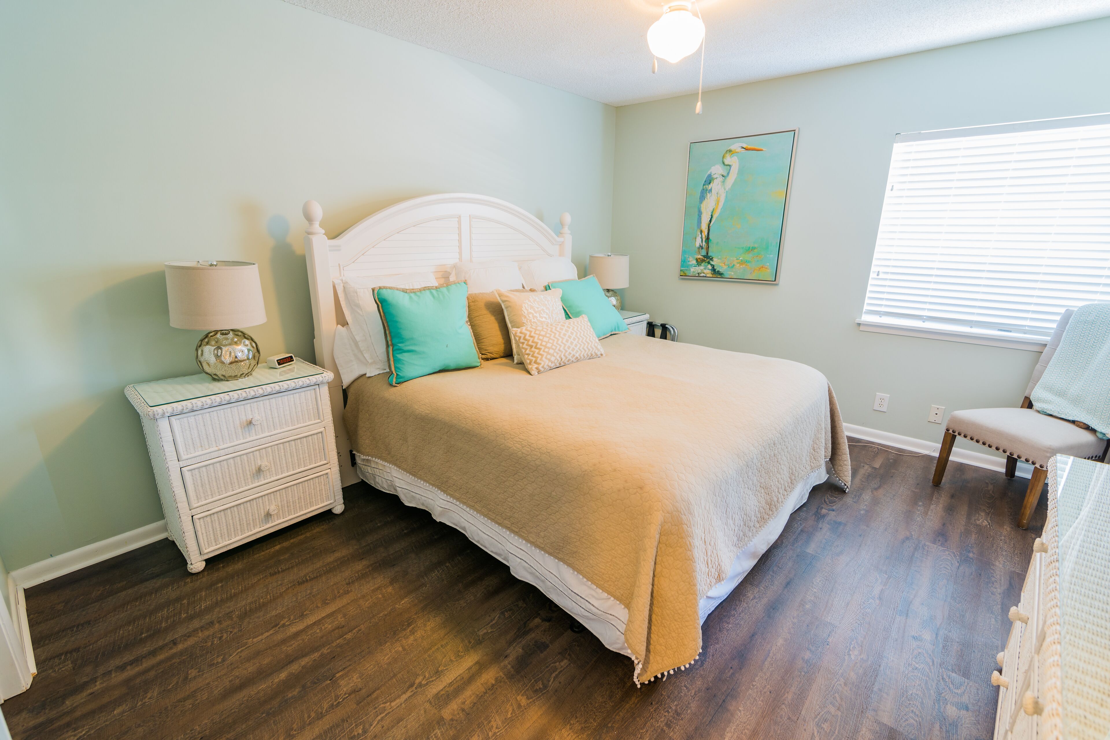 Un dormitorio con cama blanca, almohadas verde azulado y beige, una mesita de noche blanca con lámpara y suelo de madera.