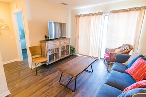 A living room with a wooden coffee table, a sofa with colorful cushions, a television on a wooden cabinet, and a wicker chair.