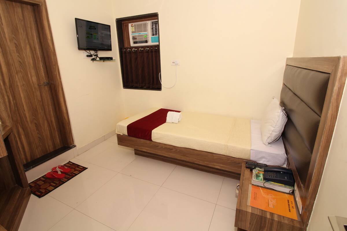 Un dormitorio pequeño y ordenado con una cama individual, una mesita de noche de madera, un televisor montado en la pared y una ventana con cortinas.