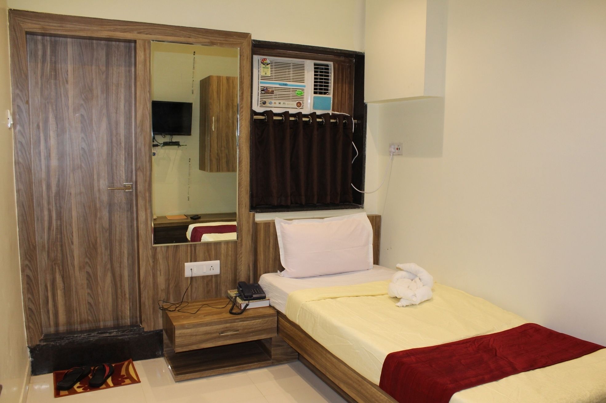 A bedroom with a wooden wardrobe, a bed with a red blanket, a bedside table with a phone and books, and a wall-mounted air conditioner.
