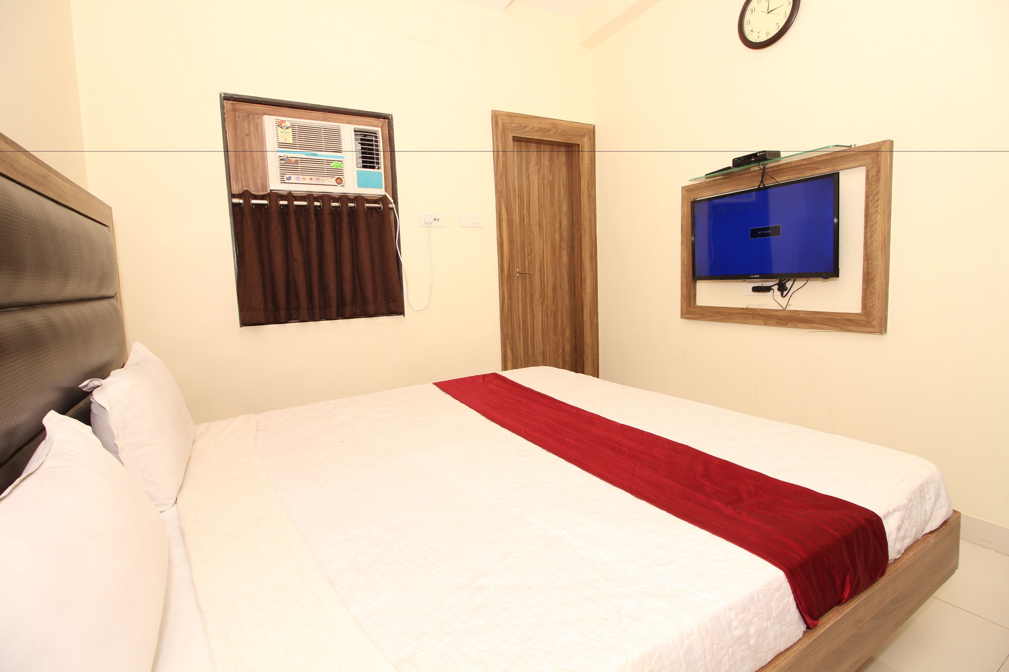 A bedroom with a bed, a window with blinds, a wooden door, and a wall-mounted TV.