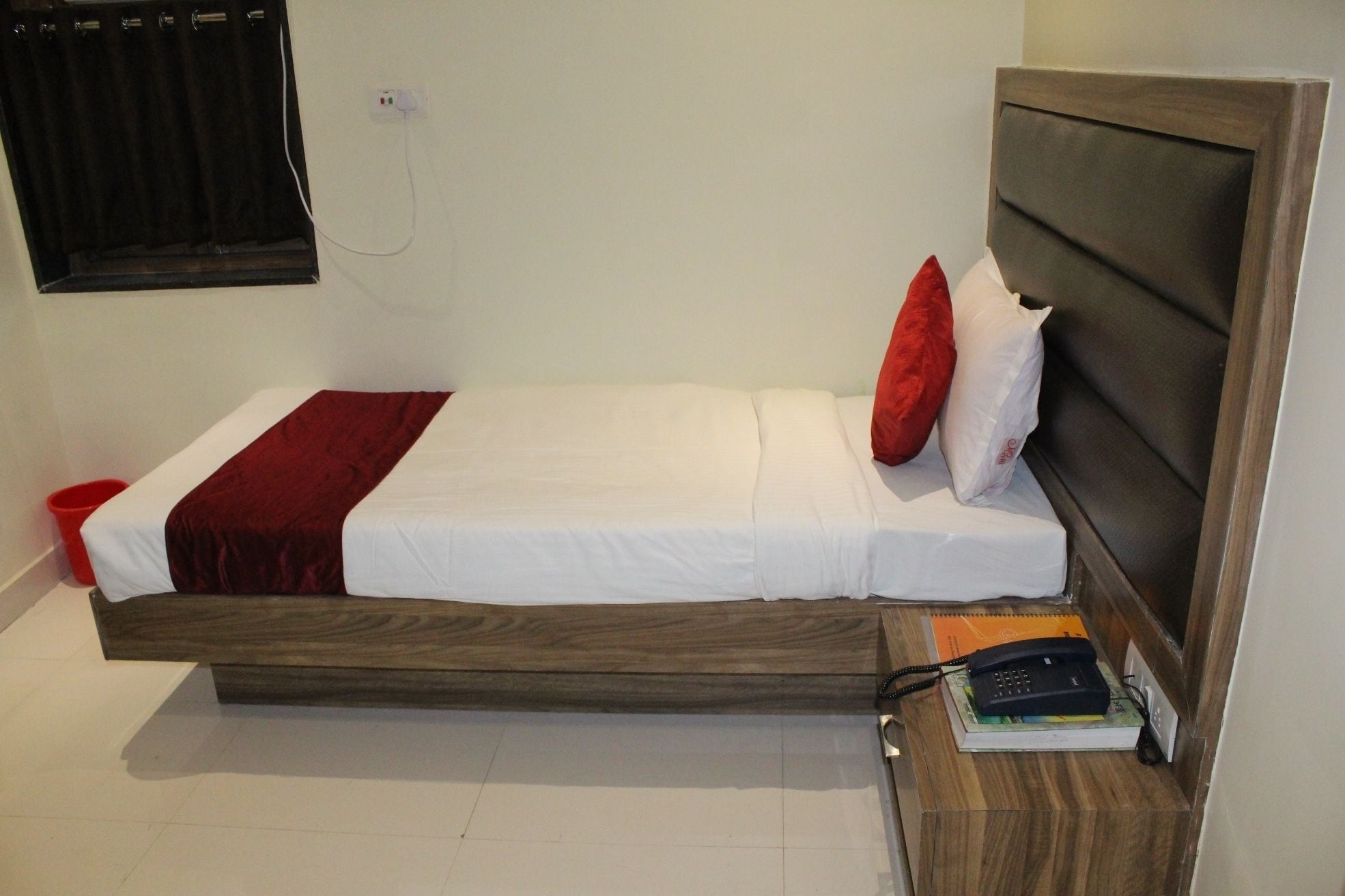 Una cama individual con cabecera de madera, ropa de cama blanca y una manta roja. Hay una mesita de noche con un teléfono y un libro.