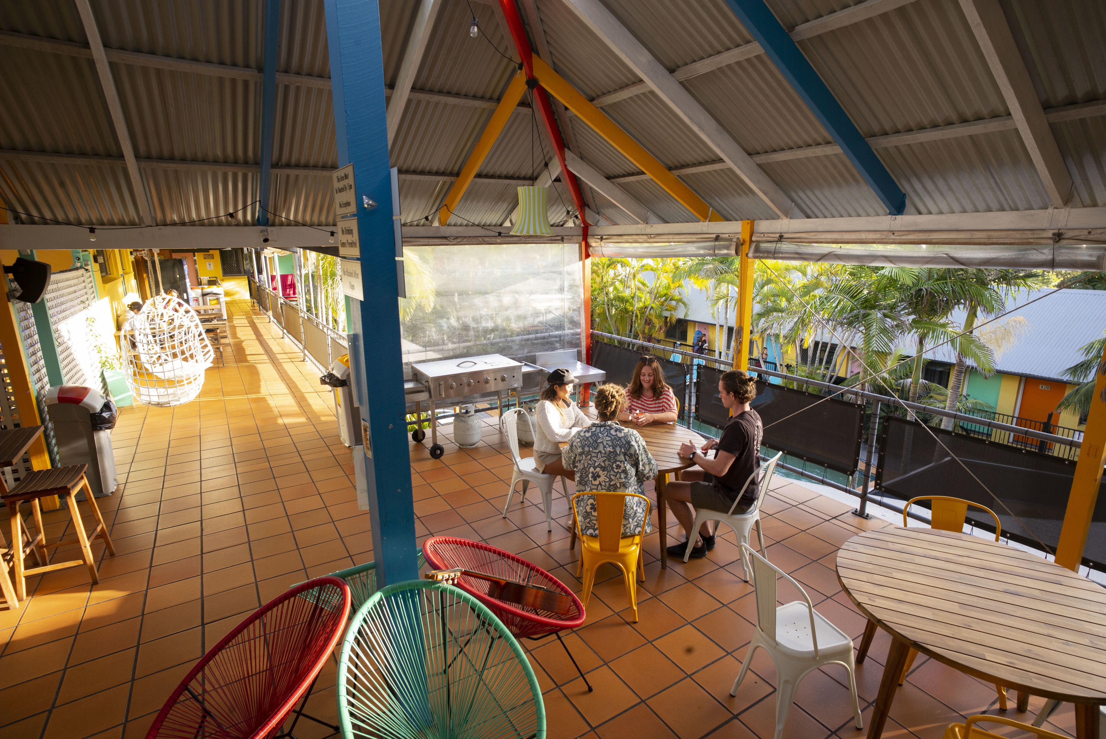 A covered outdoor seating area with tables and chairs, a grill, and a view of palm trees.