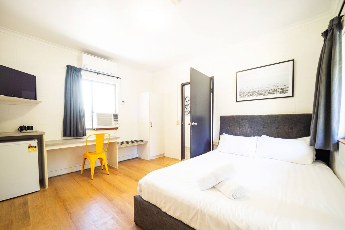 Una camera d'albergo con un letto, una scrivania, una sedia, una televisione e una finestra con tende.