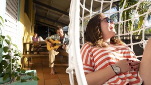 A hammock chair with a person reclining, a guitar player in the background, and a dining area with tables and chairs. - YHA Cape Byron (Byron Bay)