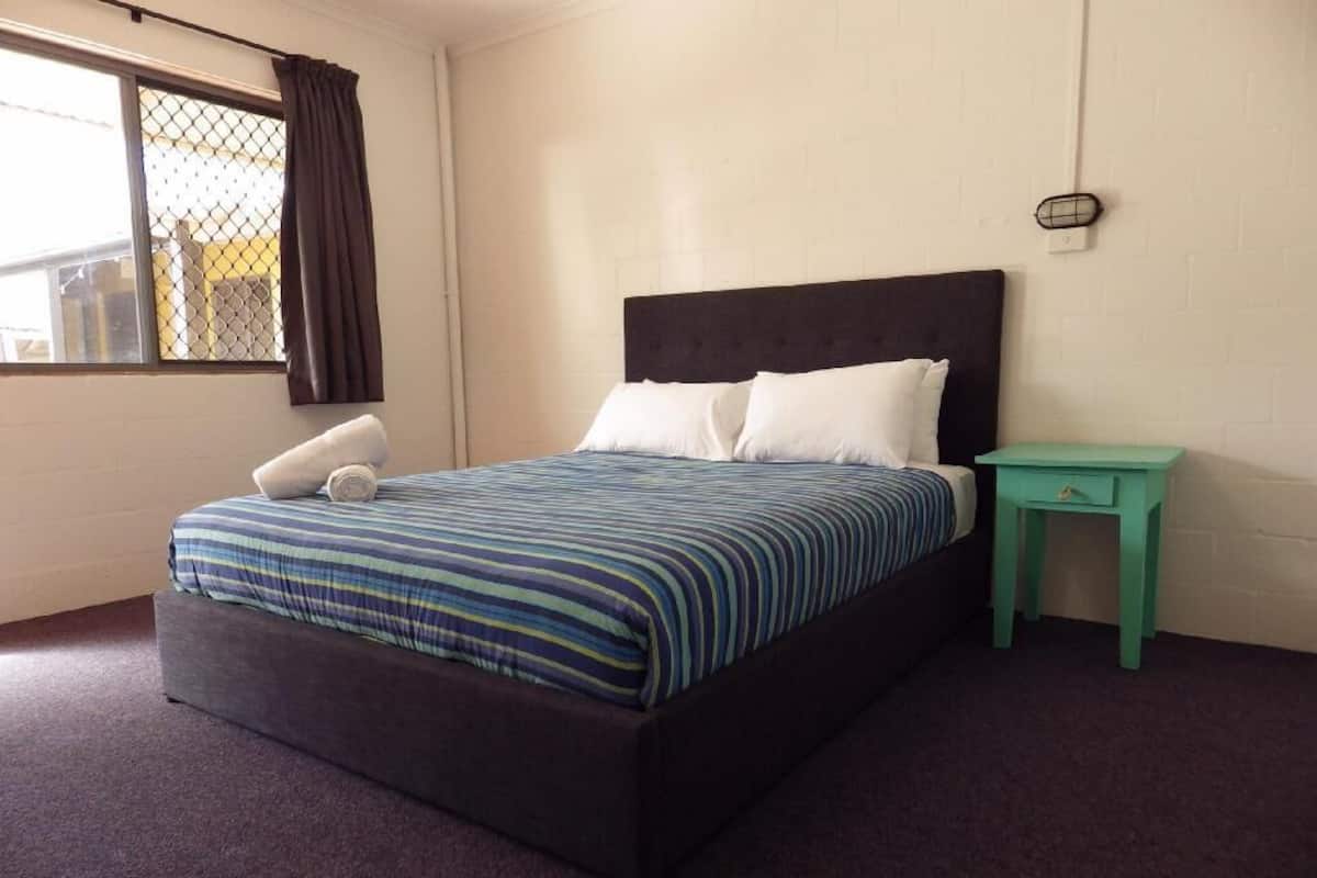 Un letto singolo con una testiera scura, un comodino piccolo verde e una finestra con tenda.
