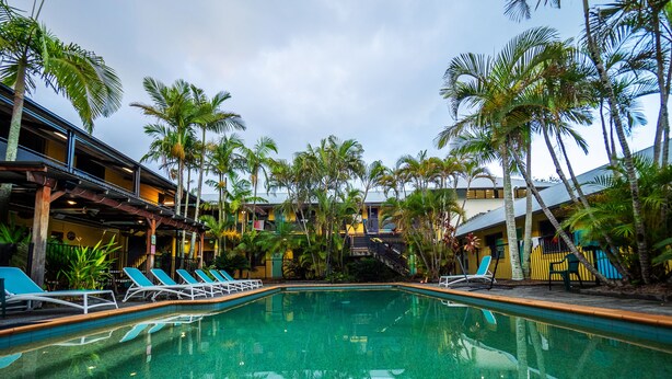 A swimming pool with lounge chairs, surrounded by palm trees and buildings with balconies. - YHA Cape Byron (Byron Bay)