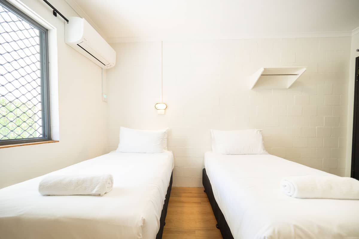 Una stanza con due letti singoli, una finestra con zanzariera, un condizionatore d'aria e un lampadario.