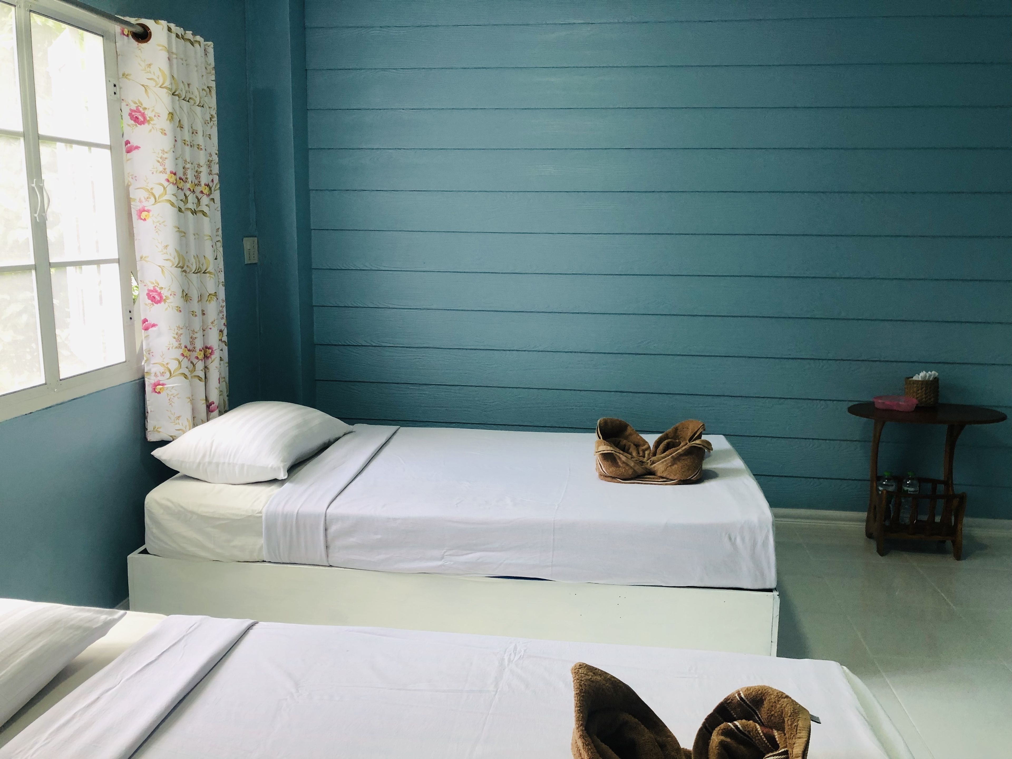 A room with two beds, a window with floral curtains, a small table with a basket, and a blue wall.