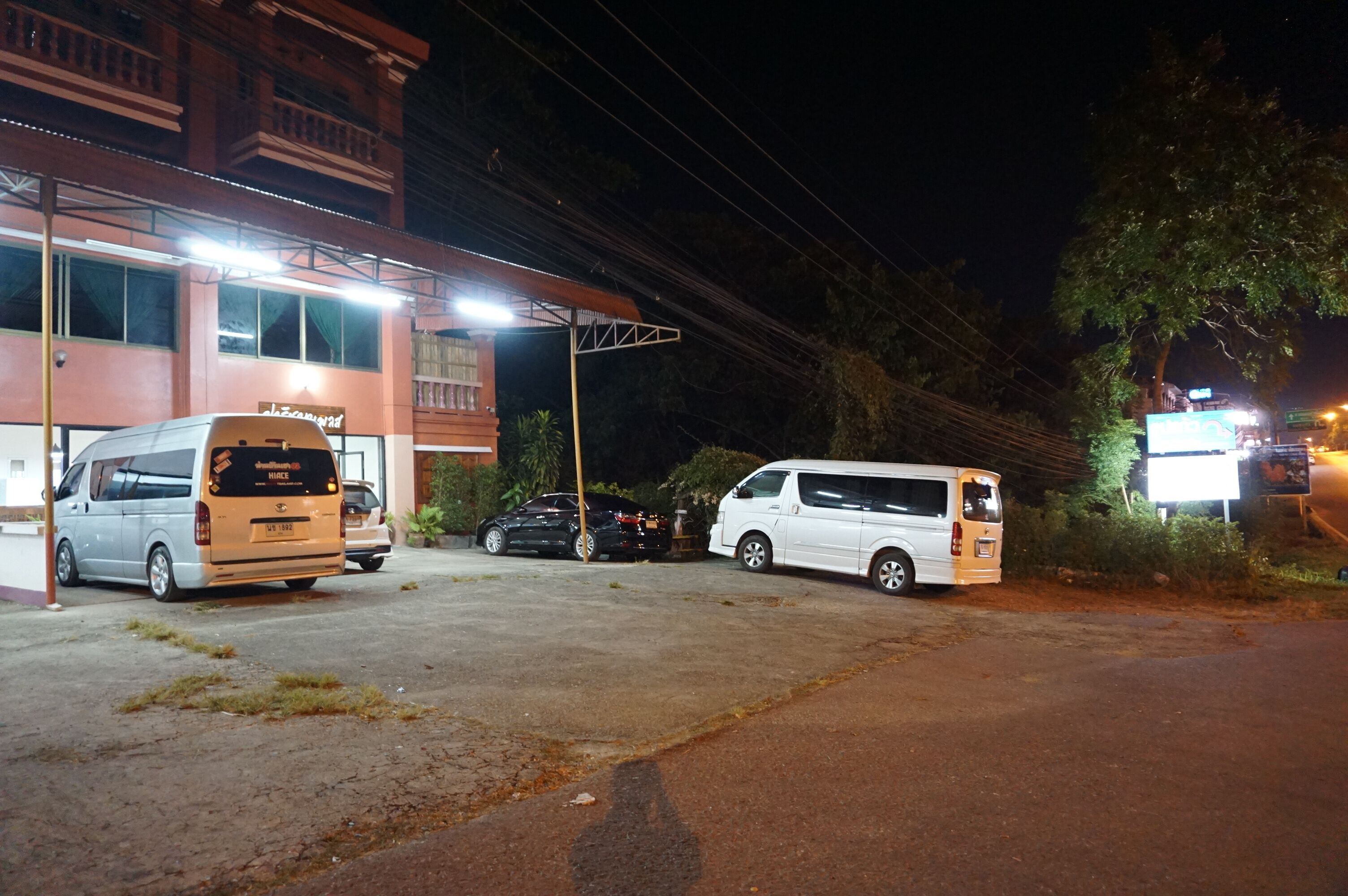 Un espace de stationnement où sont garés plusieurs véhicules, dont un fourgon blanc et une voiture noire, devant un bâtiment dont l’enseigne est illuminée.