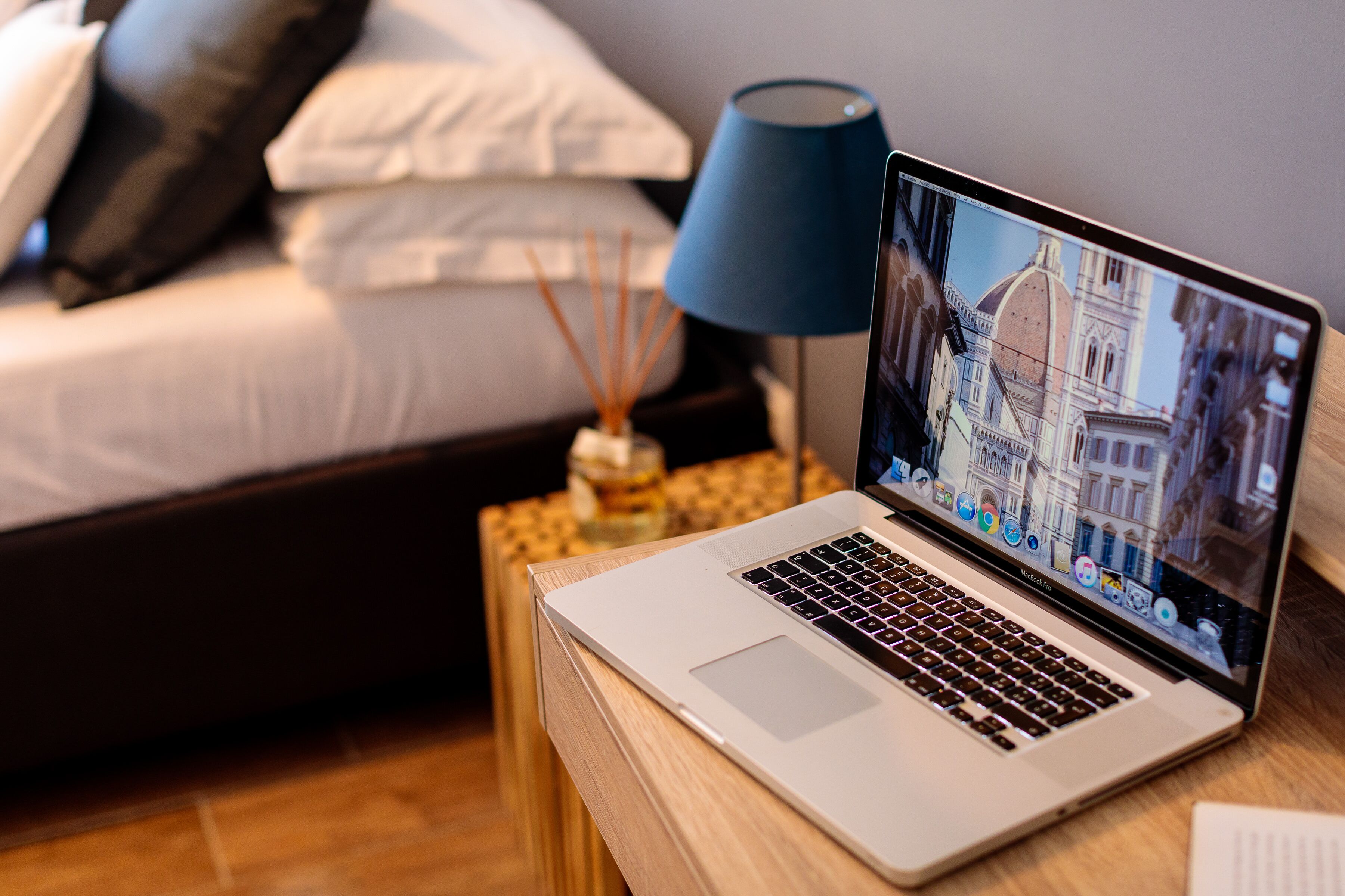 A laptop on a wooden side table with a bed in the background.