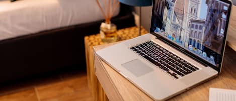 A laptop on a wooden side table with a bed in the background.