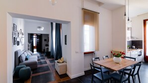 Uma sala de estar moderna com um sofá, pufes e uma mesa de jantar com cadeiras. Há uma área de cozinha com fogão e armários.