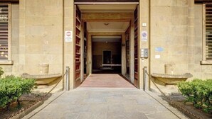 Entrada de um edifício de pedra, com um caminho central ladeado por duas pequenas fontes e uma barreira de corrente.