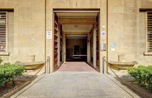 Entrada de un edificio de piedra con un camino central flanqueado por dos fuentes pequeñas y una barrera de cadena.