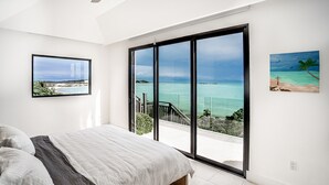 A bedroom with a large bed, a view of the ocean through a sliding glass door, and two framed pictures on the walls. - SUMMER RATE Modern Beachfront 5bed - 5bath with incredible view (providenciales)