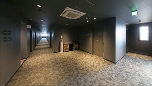 A long, narrow hallway with carpeted floors, recessed lighting, and a central air vent. - Hotel R9 Sano Fujioka (Sano)
