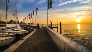 Paseo marítimo con una hilera de banderas, un muelle con barcos y un atardecer sobre el agua.