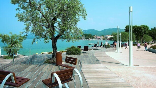 Una terraza de madera con bancos que da a un cuerpo de agua, un árbol y una vista distante de edificios.