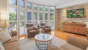 A spacious living room with large windows, a stone accent wall, a flat-screen TV, and a wooden cabinet. - #1 GUEST FAVORITE for Napili & Kapalua Bays ! (Lahaina)