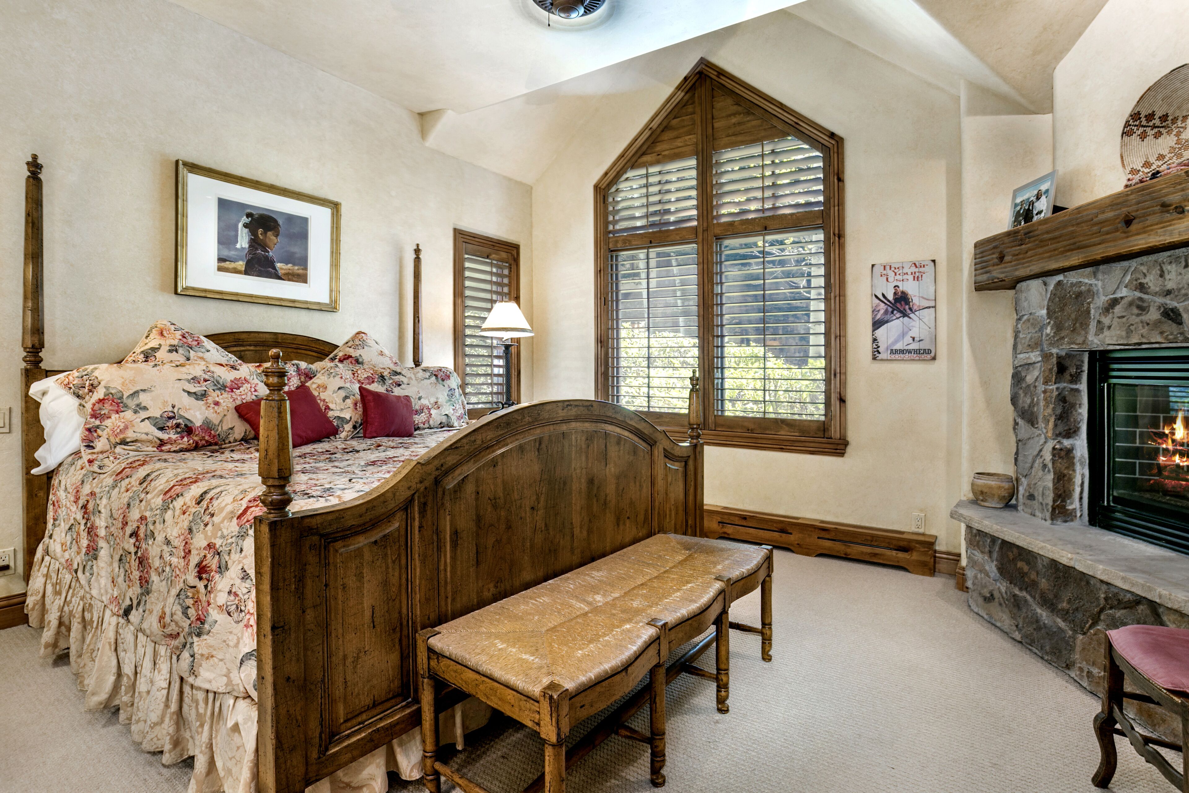 A bedroom with a wooden bed, a bench, a fireplace, and a large window with shutters.