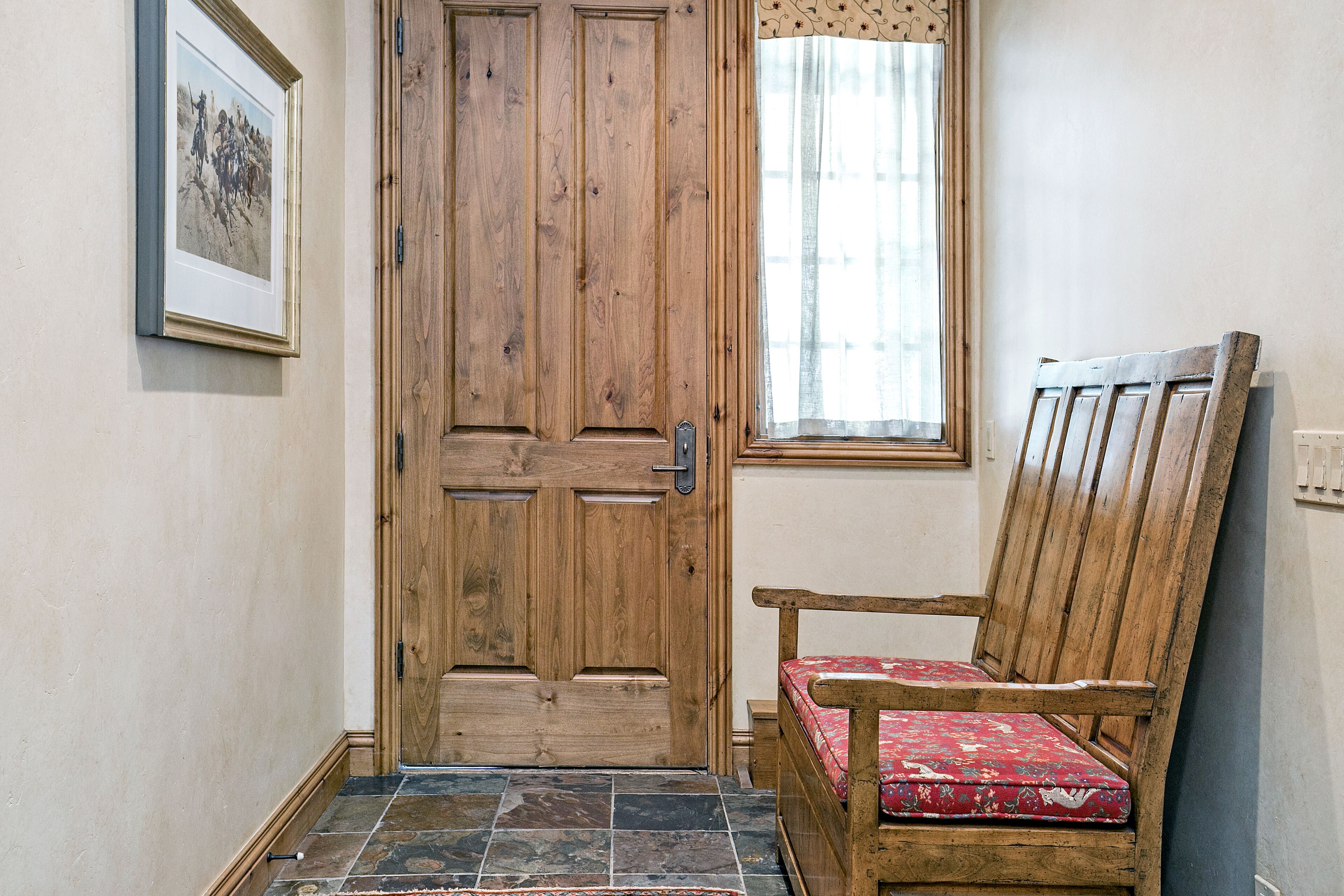 Une porte en bois avec un panneau de verre, un banc agrémenté d’un coussin à motifs et une fenêtre ornée d’un rideau léger.