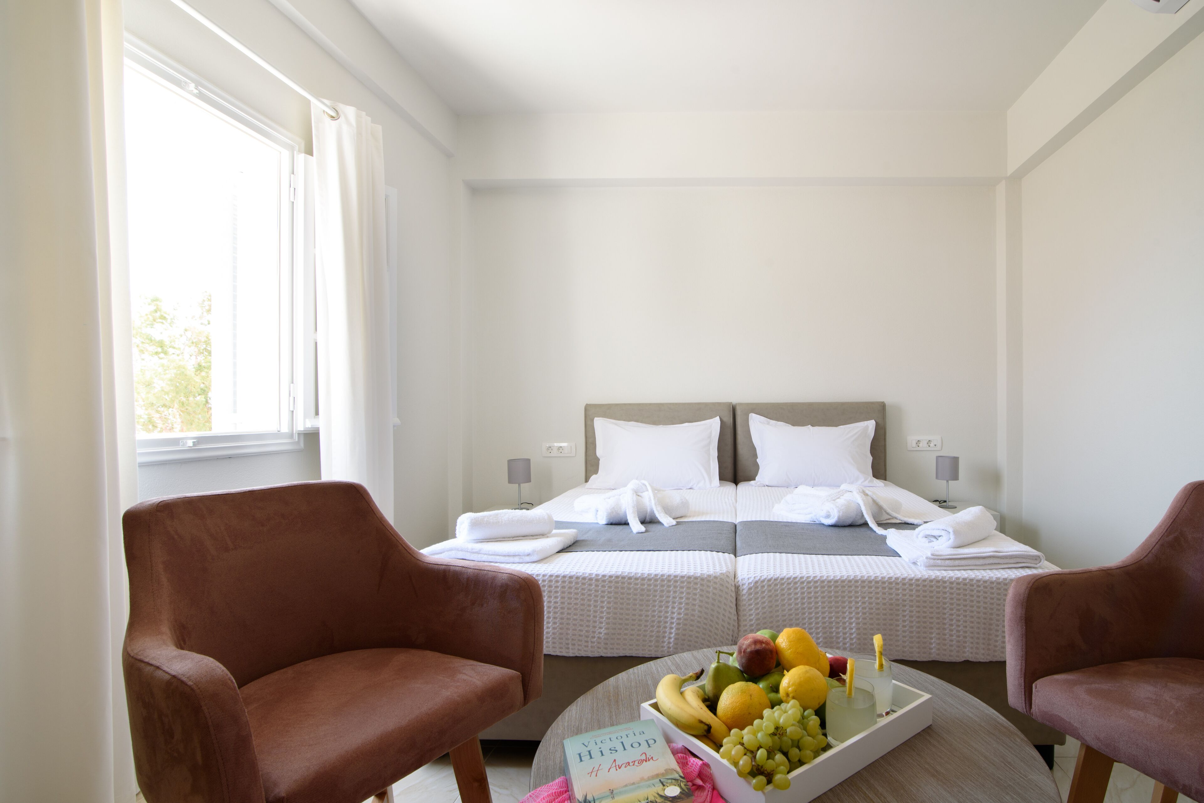 A hotel room with two beds, a round table with fruit and a book, and two chairs.