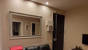 A living room with a black leather sofa, wooden flooring, a large mirror, and a flat-screen TV on a stand.