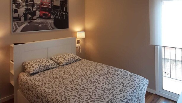 A bedroom with a large bed, a headboard, a wall-mounted lamp, and a window with a view.