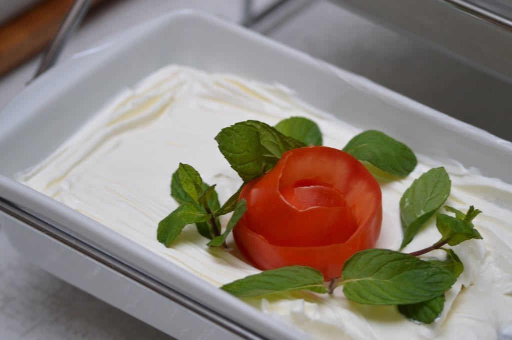 Um prato retangular branco com uma colherada de creme e um tomate esculpido em forma de rosa, decorado com folhas de hortelã.