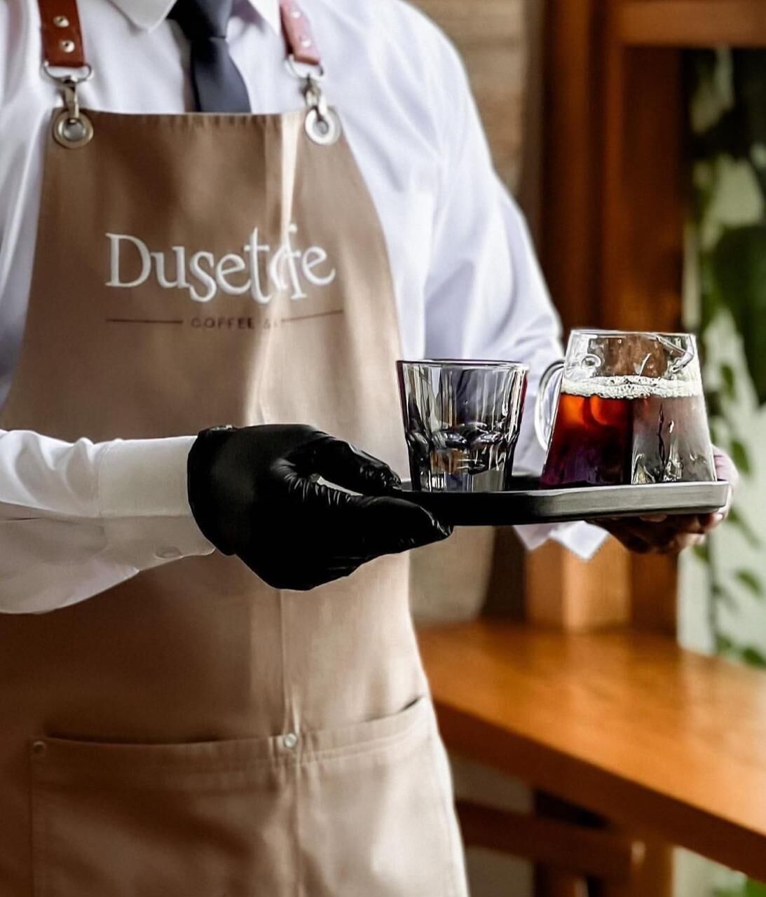 Eine Person in weißem Hemd und brauner Schürze trägt ein Tablett mit einem Glas Wasser und einem Glas Eistee.