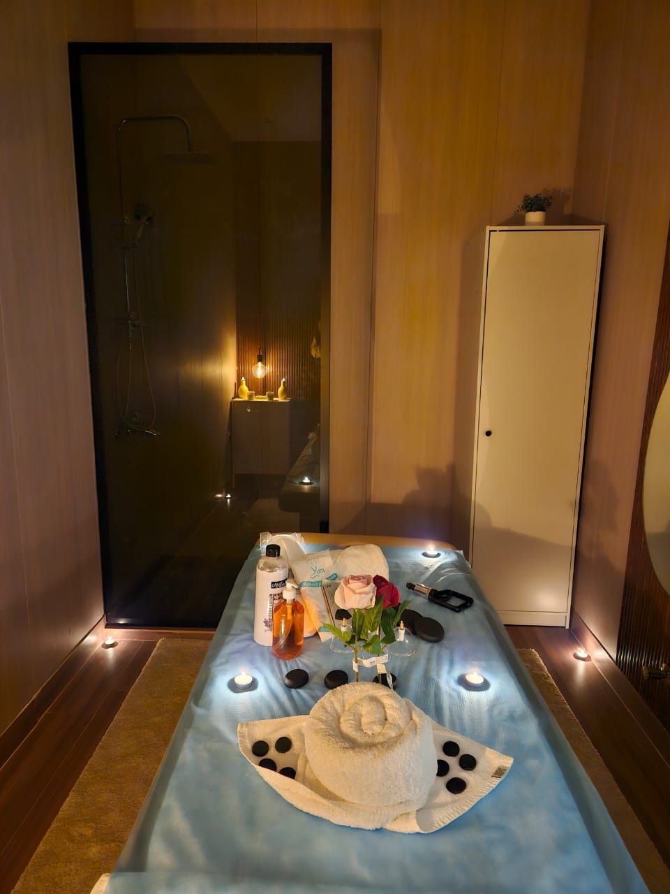 Habitación de hotel con una camilla de masaje, una mesita de noche con artículos de aseo, un armario y la puerta a un área de ducha.