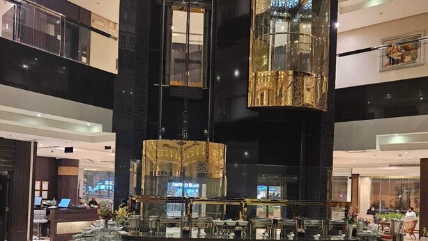 Un interior moderno con una barra central, varias mesas de comedor con cojines verdes y un techo alto con superficies reflectantes.