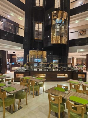 A modern interior with a central bar area, multiple dining tables with green cushions, and a high ceiling with reflective surfaces. - Duset Suites (Riyadh)
