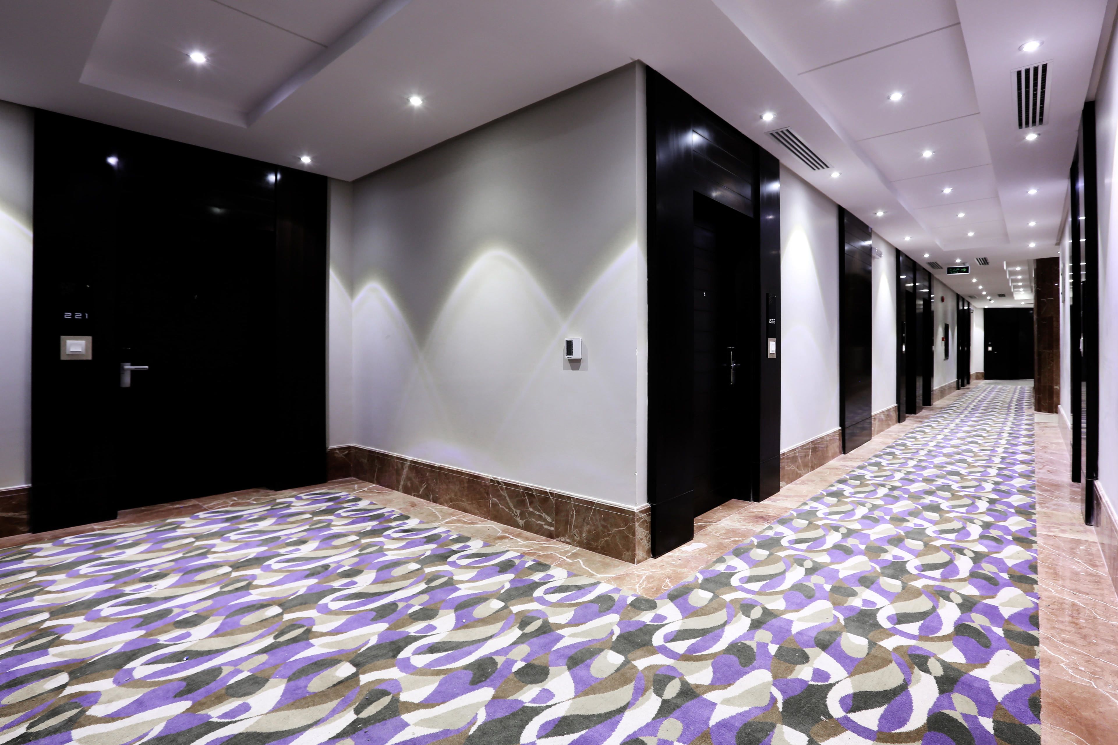 A hotel corridor with patterned carpet, black doors, and recessed lighting.