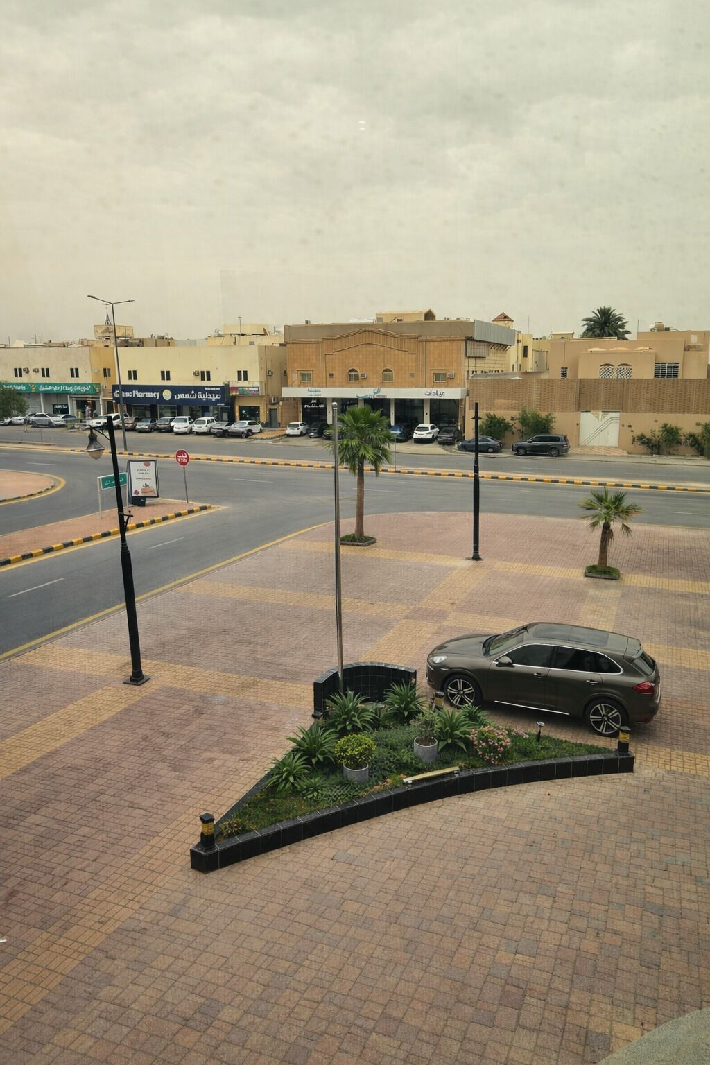 A paved pedestrian area with a car parked on the side, a roundabout, and buildings with signage in a foreign language.