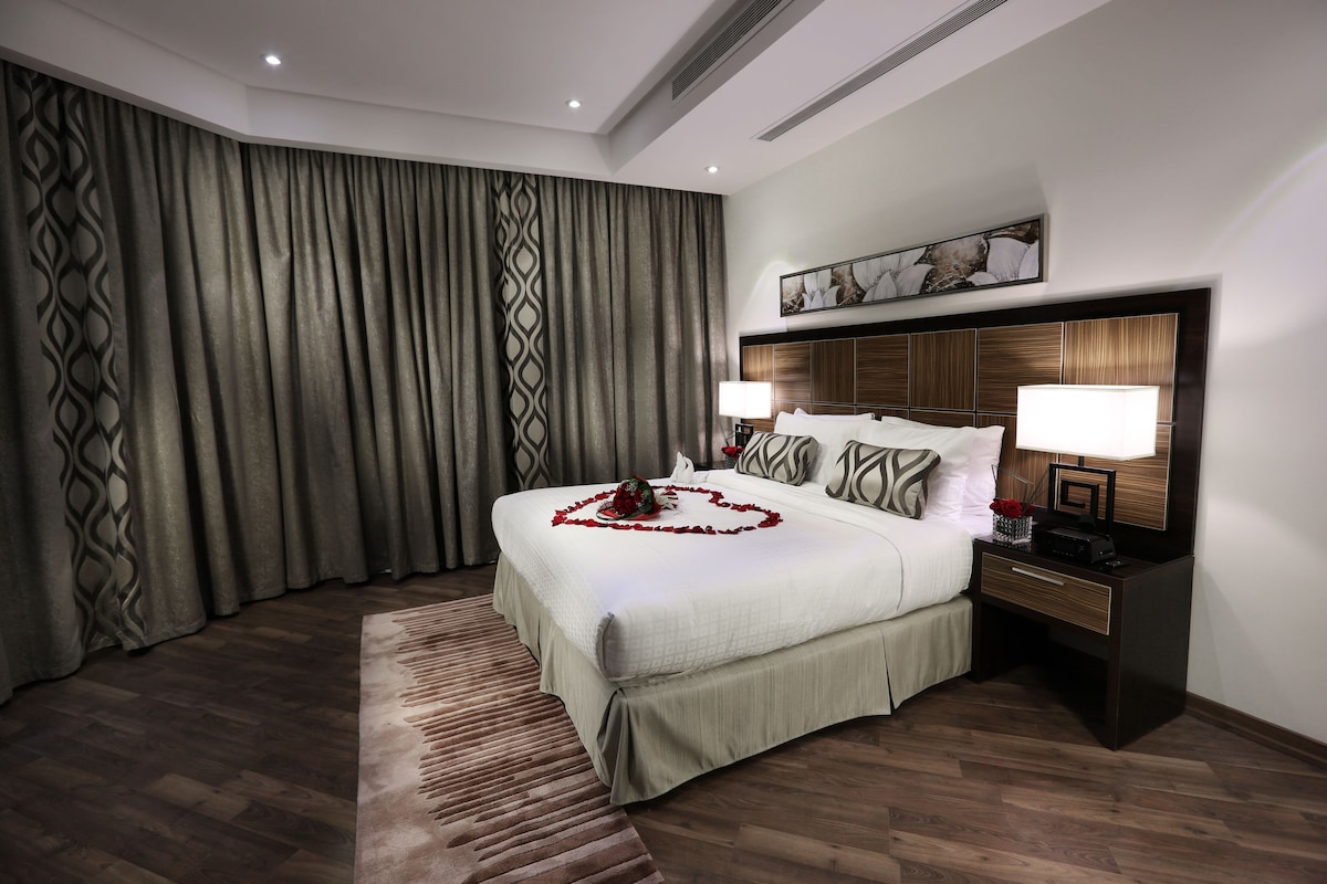 Una habitación de hotel con una cama grande, una mesita de noche con lámpara y un cabecero con panel decorativo.