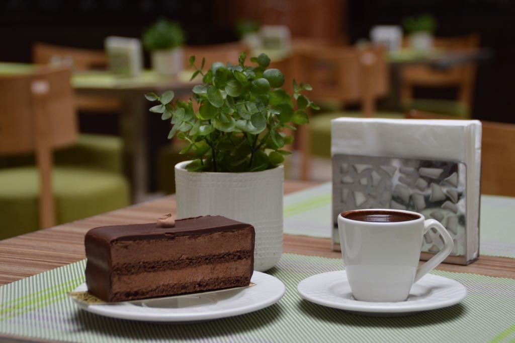 A slice of chocolate cake on a plate, a white cup of coffee, and a potted plant on a table.