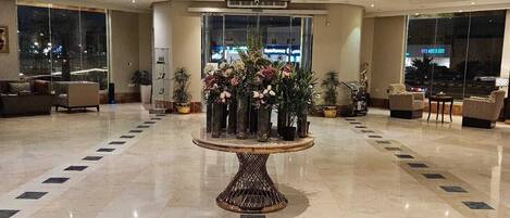 Un vestíbulo amplio con un arreglo floral central sobre una mesa redonda, ventanas grandes y áreas de descanso.