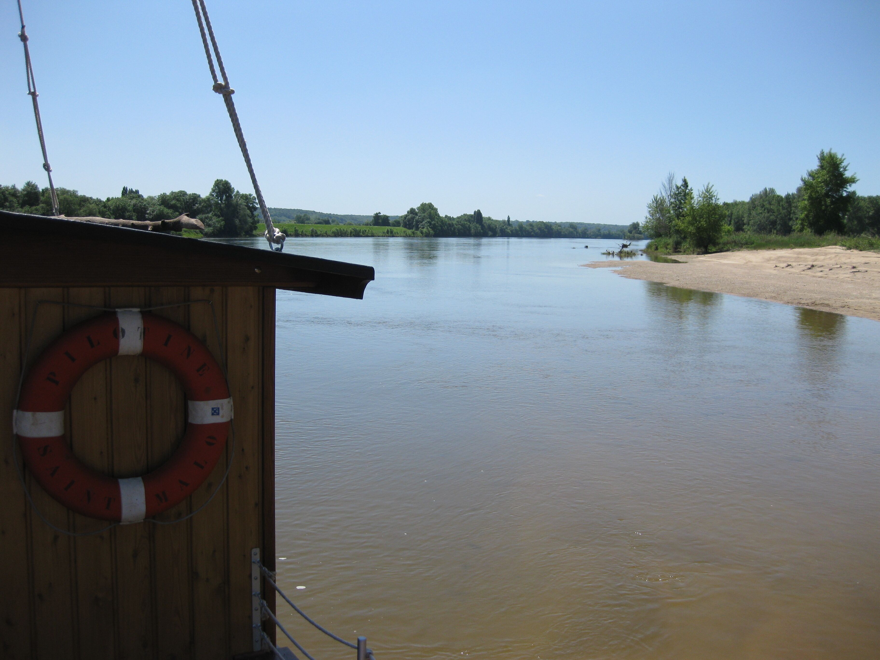 Een houten constructie met een reddingsboei, uitkijkend over een kalme rivier met een zandige oever en begroening op de achtergrond.