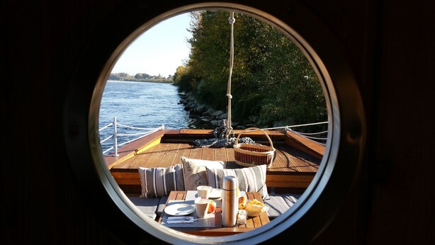 Une vue à travers une fenêtre circulaire sur le pont d’un bateau, avec une table dressée pour le petit-déjeuner, comprenant un panier, un thermos et un pain.
