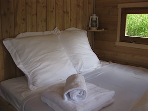 Une pièce de cabane en bois comprenant un lit, des oreillers, une serviette enroulée, une lanterne et une fenêtre donnant sur un paysage verdoyant.