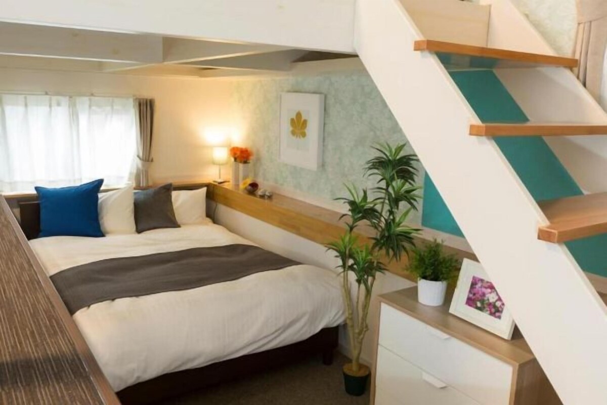 A bedroom with a bed, bedside tables, a desk, a chair, a potted plant, and a staircase.