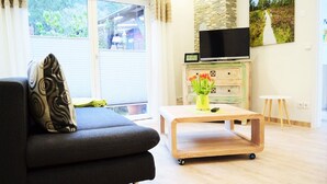 Ein Wohnzimmer mit einer Couch, einem Holztisch mit Blumenvase, einem Fernseher auf einer Kommode und Blick ins Grüne.