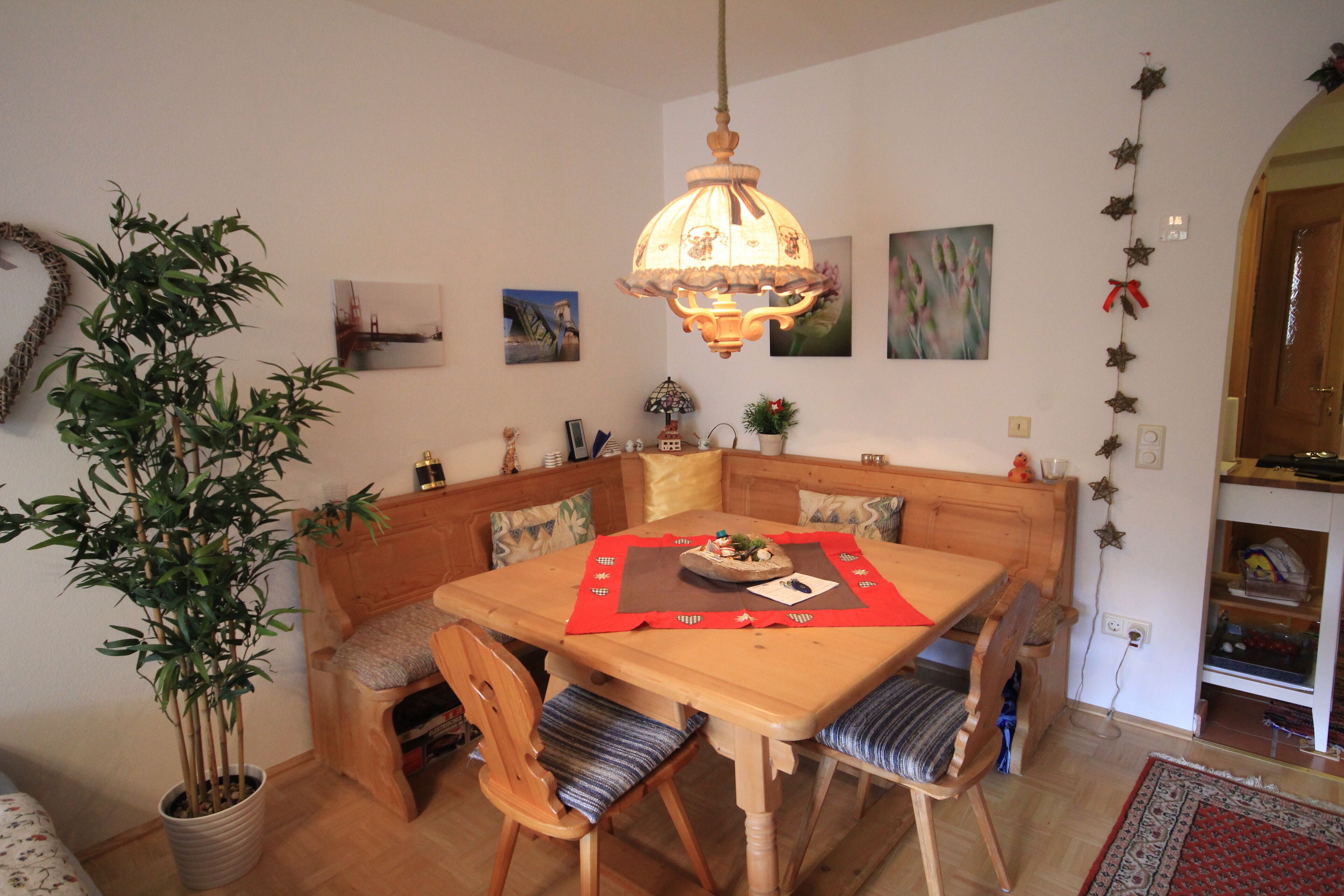 A dining area with a wooden table, chairs, and a hanging light fixture. There is a potted plant and artwork on the walls.