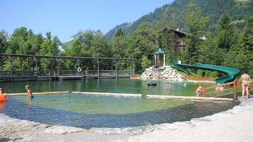 A swimming pool with a wooden deck and a green slide, surrounded by trees and a mountain in the background.