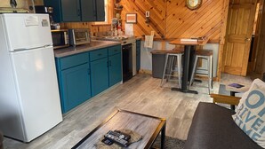 Una cocina compacta con armarios color turquesa, un refrigerador blanco y una cafetera. Hay un comedor de madera con taburetes y una mesa, y un sofá con un cojín que dice "DRIVE".